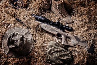 A camouflaged sniper rifle lies amidst a backdrop of tactical gear and a ghillie suit. Surrounding the rifle are a pair of tan combat boots, a camouflage hat, and other military accessories. The colors and textures blend with the natural environment, indicating a setup for concealment in an outdoor setting.