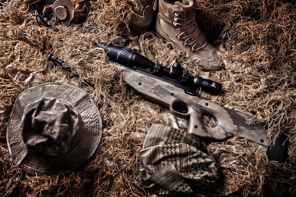 A camouflaged sniper rifle lies amidst a backdrop of tactical gear and a ghillie suit. Surrounding the rifle are a pair of tan combat boots, a camouflage hat, and other military accessories. The colors and textures blend with the natural environment, indicating a setup for concealment in an outdoor setting.