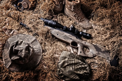 A camouflaged sniper rifle lies amidst a backdrop of tactical gear and a ghillie suit. Surrounding the rifle are a pair of tan combat boots, a camouflage hat, and other military accessories. The colors and textures blend with the natural environment, indicating a setup for concealment in an outdoor setting.