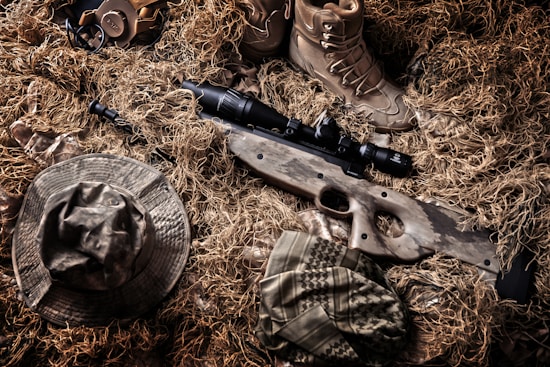 A camouflaged sniper rifle lies amidst a backdrop of tactical gear and a ghillie suit. Surrounding the rifle are a pair of tan combat boots, a camouflage hat, and other military accessories. The colors and textures blend with the natural environment, indicating a setup for concealment in an outdoor setting.