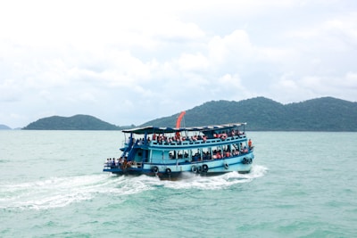 A blue ferry boat filled with passengers cruises across a calm turquoise sea. The boat is adorned with orange life jackets hanging around the deck. In the background, lush green islands rise from the water under a cloudy sky.
