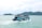 A blue ferry boat filled with passengers cruises across a calm turquoise sea. The boat is adorned with orange life jackets hanging around the deck. In the background, lush green islands rise from the water under a cloudy sky.