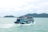 A blue ferry boat filled with passengers cruises across a calm turquoise sea. The boat is adorned with orange life jackets hanging around the deck. In the background, lush green islands rise from the water under a cloudy sky.