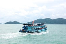 A blue ferry boat filled with passengers cruises across a calm turquoise sea. The boat is adorned with orange life jackets hanging around the deck. In the background, lush green islands rise from the water under a cloudy sky.
