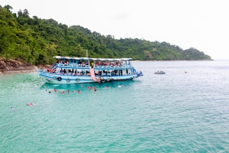 A large blue and white boat filled with numerous people floats on calm turquoise waters, with a lush green forested island as the backdrop. Several people are swimming around the boat, enjoying the clear water. A smaller boat can be seen in the background.