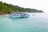 A large blue and white boat filled with numerous people floats on calm turquoise waters, with a lush green forested island as the backdrop. Several people are swimming around the boat, enjoying the clear water. A smaller boat can be seen in the background.