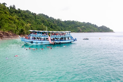 A large blue and white boat filled with numerous people floats on calm turquoise waters, with a lush green forested island as the backdrop. Several people are swimming around the boat, enjoying the clear water. A smaller boat can be seen in the background.