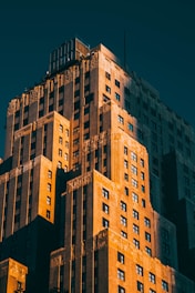 A tall, multi-layered Art Deco building is captured in warm, late afternoon sunlight, emphasizing its intricate architectural details and vertical lines. The sky is a deep, contrasting teal, enhancing the building's orange and brown hues.