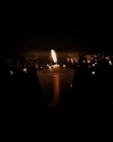 A single lit candle is placed in a transparent holder against a dark background. The candle's flame gently illuminates its immediate surroundings. In the distance, numerous small lights create a bokeh effect, suggesting a gathering or vigil.
