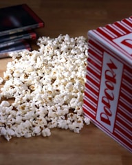 A pile of popcorn spilled out from a red and white striped container, with several DVD cases laying nearby on a wooden surface.