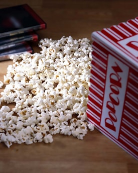 A pile of popcorn spilled out from a red and white striped container, with several DVD cases laying nearby on a wooden surface.