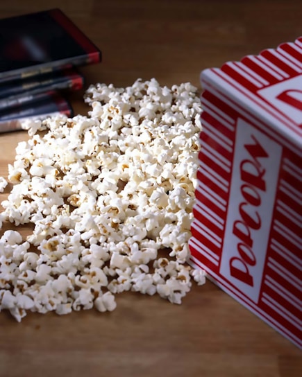 A pile of popcorn spilled out from a red and white striped container, with several DVD cases laying nearby on a wooden surface.