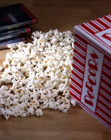 A pile of popcorn spilled out from a red and white striped container, with several DVD cases laying nearby on a wooden surface.