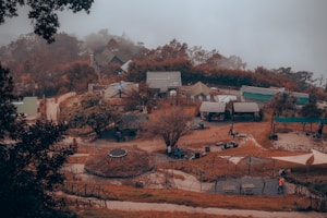 An elevated view of a small village or campsite surrounded by lush trees and vegetation. There are several tents and small structures scattered around, with pathways connecting them. The autumnal hues suggest a chilly, foggy day as mist envelops the background. A few people are visible, engaged in various activities.