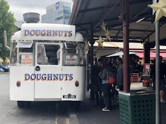 A white food truck with large, colorful signage advertising doughnuts is parked in an urban area. The truck has signs indicating hot doughnuts are now ready. People are gathered around the truck, likely waiting to make a purchase. The surrounding area includes a covered market or street with some small stalls and decorations like hanging stars.