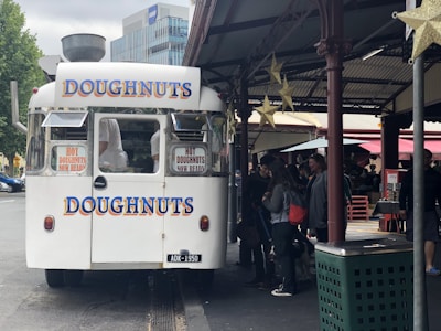 A white food truck with large, colorful signage advertising doughnuts is parked in an urban area. The truck has signs indicating hot doughnuts are now ready. People are gathered around the truck, likely waiting to make a purchase. The surrounding area includes a covered market or street with some small stalls and decorations like hanging stars.