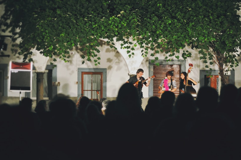A small musical group is performing on an outdoor stage in front of a crowd. The backdrop includes a building with arched windows and trailing greenery above the performers' heads. The performers are playing string instruments, and the scene is illuminated under bright stage lights.