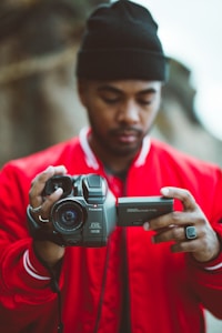 A person wearing a red jacket and a black beanie is holding a Panasonic video camera, examining the screen. The focus is on the camera, with the person slightly blurred in the background, creating a depth of field effect.