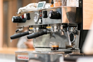 A close-up of an espresso machine in action, with robust metal handles and a digital display. It features two portafilters and a single shot of espresso being brewed into a small glass measuring cup.