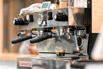 A close-up of an espresso machine in action, with robust metal handles and a digital display. It features two portafilters and a single shot of espresso being brewed into a small glass measuring cup.
