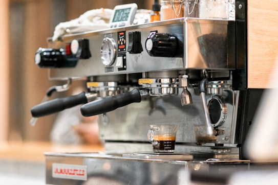 A close-up of an espresso machine in action, with robust metal handles and a digital display. It features two portafilters and a single shot of espresso being brewed into a small glass measuring cup.