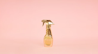 A golden spray bottle stands upright against a soft pink background, creating a striking contrast with its metallic shine.