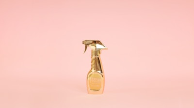 A golden spray bottle stands upright against a soft pink background, creating a striking contrast with its metallic shine.