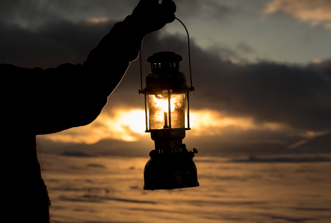 person standing on seashore and holding kerosene lantern lamp during sunset
