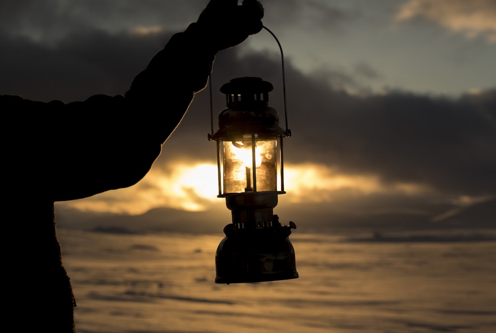 person standing on seashore and holding kerosene lantern lamp during sunset