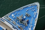 The image shows the bow of a large ship, viewed from above, featuring a deck painted in blue. Various nautical equipment such as winches, chains, ropes, and an anchor are visible. The ship is situated in calm green water, and the deck has a neat, organized appearance with multiple white bollards and mooring stations aligned along the edges.