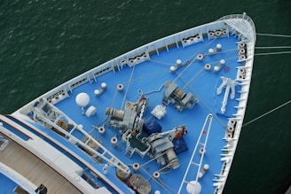 The image shows the bow of a large ship, viewed from above, featuring a deck painted in blue. Various nautical equipment such as winches, chains, ropes, and an anchor are visible. The ship is situated in calm green water, and the deck has a neat, organized appearance with multiple white bollards and mooring stations aligned along the edges.