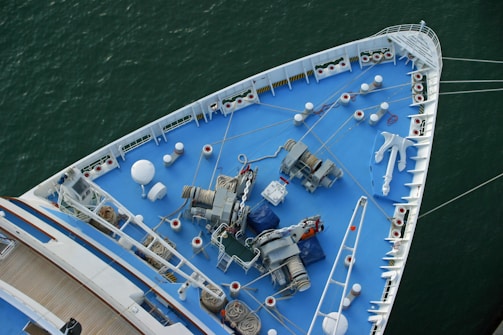 The image shows the bow of a large ship, viewed from above, featuring a deck painted in blue. Various nautical equipment such as winches, chains, ropes, and an anchor are visible. The ship is situated in calm green water, and the deck has a neat, organized appearance with multiple white bollards and mooring stations aligned along the edges.