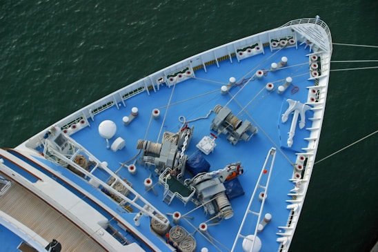 The image shows the bow of a large ship, viewed from above, featuring a deck painted in blue. Various nautical equipment such as winches, chains, ropes, and an anchor are visible. The ship is situated in calm green water, and the deck has a neat, organized appearance with multiple white bollards and mooring stations aligned along the edges.