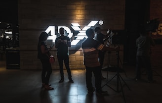 Several people stand silhouetted against a brightly lit sign that reads '4DX' on a brick wall. The individuals are casually dressed, with some appearing to interact with electronic devices, while others seem to be working with camera equipment.