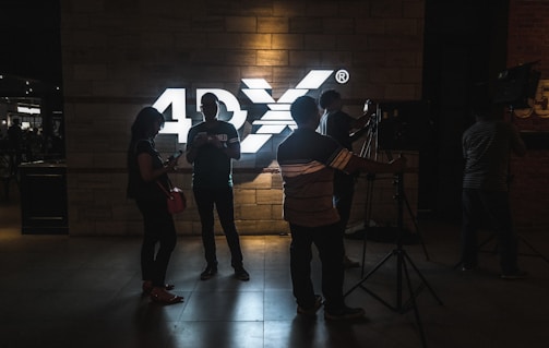 Several people stand silhouetted against a brightly lit sign that reads '4DX' on a brick wall. The individuals are casually dressed, with some appearing to interact with electronic devices, while others seem to be working with camera equipment.