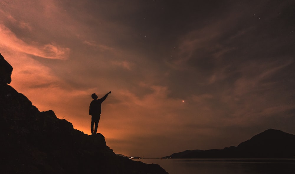 Silhouettenfotografie einer Person, die auf einem Felsen neben einem Gewässer steht