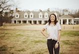 A young woman stands smiling in the foreground with her hands on her hips. She is wearing a light pink T-shirt, dark jeans, a watch, and jewelry. Behind her is a large, elegant house with multiple dormer windows and a broad porch. The setting is outdoors with a grassy area and some trees in the background.