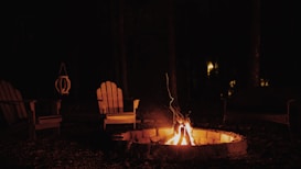 A cozy outdoor setting featuring a stone fire pit glowing in the dark. Surrounding the fire pit are wooden Adirondack chairs, positioned for relaxation. The background is enveloped in darkness with faint, ambient lights visible among the trees, adding depth to the tranquil night scene.