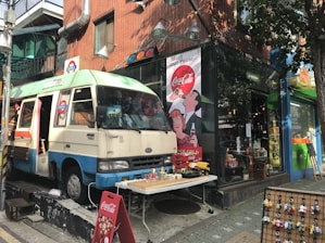 A vintage-style street scene featuring a parked retro van with a weathered exterior, situated next to a small shop. The van is decorated with faded colorful paint and has various items displayed on a table in front of it, including small toys and a Coca-Cola sign. The shop window displays creative merchandise, and there's a nostalgic Coca-Cola poster on the brick wall. Additional keychains and toys are organized on a rack by the sidewalk.