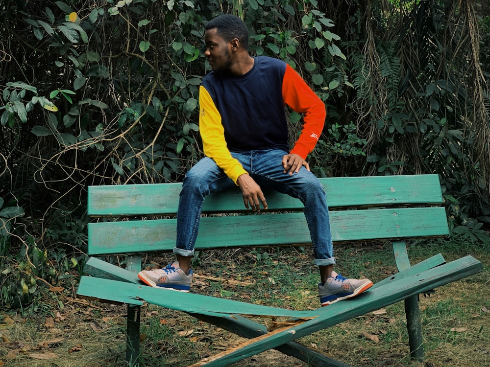 man in yellow, red, and black long-sleeved shirt and blue jeans sitting on wrecked bench backrest