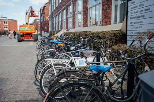 A row of bicycles is parked outside a building with brick walls and many windows. In the background, a orange construction lift vehicle is visible. There are people walking and a sign with Swedish text is placed next to the bicycles.