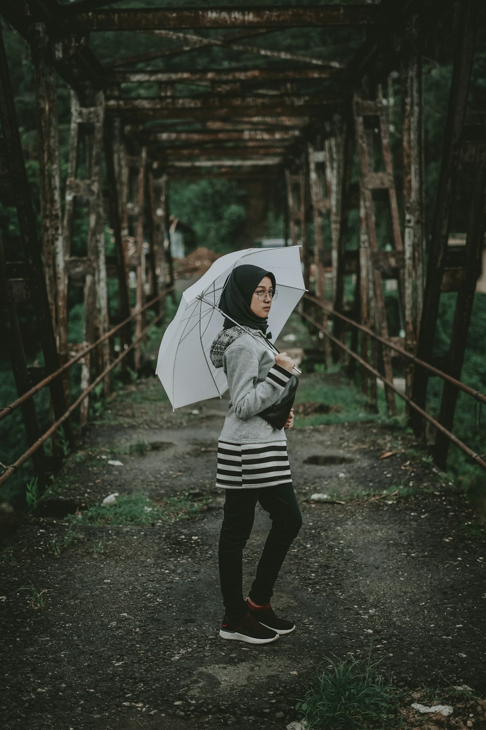 woman wearing black hijab standing on abandoned bridge and holding umbrella