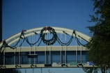 A bridge adorned with garlands and bows, seen against a clear blue sky. The garlands are arranged symmetrically with festive red bows. Traffic signs and green lights are visible below the bridge, indicating road names and directions. Trees and urban elements frame the structure.