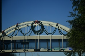 A bridge adorned with garlands and bows, seen against a clear blue sky. The garlands are arranged symmetrically with festive red bows. Traffic signs and green lights are visible below the bridge, indicating road names and directions. Trees and urban elements frame the structure.