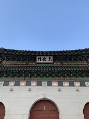 A traditional Korean architectural structure featuring intricate wooden designs and vibrant colors, including red, green, and blue elements. A large sign with Chinese characters is prominently displayed at the center, above a stone and wooden entrance.