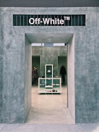 The entrance to a modern retail store with a minimalist design features a grey textured facade. The interior is visible through the open doorway, displaying a clean, organized layout with shelves holding various fashion items. A mannequin with a colorful dress and a coat rack are situated inside.