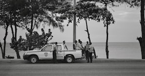 Several people wearing uniforms are gathered around a parked pickup truck on a road by the ocean. The background features trees with sparse leaves, and the sea is visible beyond the edge of the road.