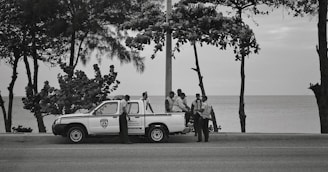 Several people wearing uniforms are gathered around a parked pickup truck on a road by the ocean. The background features trees with sparse leaves, and the sea is visible beyond the edge of the road.
