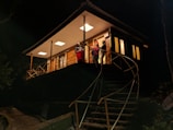 A modern-looking house with a triangular roof is elevated on stilts, accessed by a winding staircase. The house is warmly lit from within, suggesting a cozy interior. Several people are standing on the balcony, appearing to be engaged in conversation. The surroundings are dark, indicating it is nighttime.