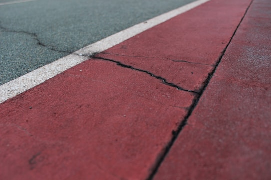 A close-up view of a cracked running track or sports field with a red surface and a dividing white line. The surface appears worn and aged, with visible texture and signs of use.